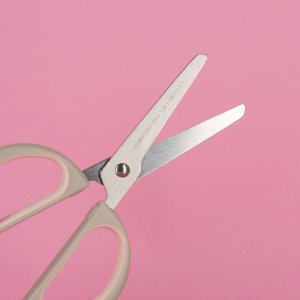 A close-up shot of a pair of grey handled scissors with a cat theme. The blades are partially open and displayed on a pink background. The handles are a light grey with pale pink inner grips The metal blades have some text on them and the pivot screw point is visible.