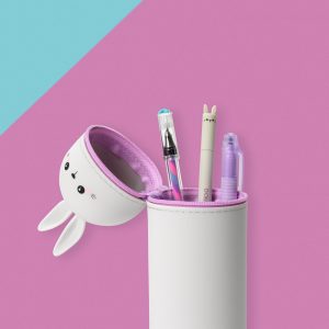 A shot of a cute, grey, bunny-shaped pencil case by Legami, set on a multi-colored background of light blue and pink. The pencil case is standing upright with its zipped lid open. The lid, which forms the bunny's head, is shown dangling at the side with a kawaii-style face. The open pencil case reveals a selection of stationery inside, including a pen with a bunny topper.