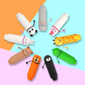 A creative product shot displaying a range of nine different animal-themed pencil cases by Legami, arranged in a circular pattern on a colorful background of blue, orange and pink. The pencil cases are, starting at the top and going around in a clockwise direction: a grey cat, a white unicorn, a yellow giraffe with orange spots, a green dinosaur, a black penguin, an orange tiger, a brown teddy bear, a grey bunny and a white panda. All of the pencil cases are lying down and have a cute, kawaii-style face.