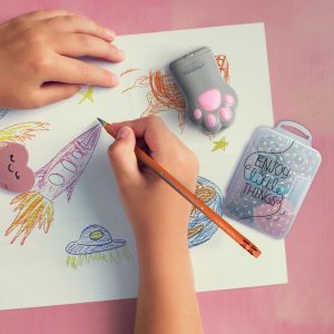 A pair of hands is shown drawing on a piece of paper with a pencil. The drawing includes a rocket ship, a planet, and other space-themed objects. A grey, cat paw-shaped pencil sharpener is sitting on the paper, next to a clear plastic box filled with small stationery items. The surface underneath the paper is a dusty pink colour.