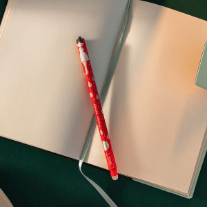 A red Legami erasable pen, featuring a ballerina design on its cap and "Jingle Dance" written vertically along its body, rests diagonally across an open notebook. The notebook has cream-colored blank pages and a light green ribbon bookmark lying across the left page. The notebook's cover is also light green, and the surface it rests on appears to be a dark green colour. The lighting casts a soft shadow from the pen onto the page, creating a gentle atmosphere.