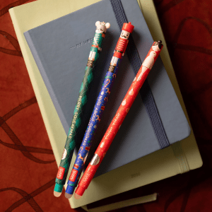 Three Christmas-themed erasable pens are laid diagonally across a stack of two notebooks. The top notebook is a dark grey-blue with an elastic band and "MY NOTEBOOK" debossed near the top. The bottom notebook is a lighter sage green. The background is a dark reddish-brown surface with a subtle texture or pattern. From top left to bottom right, the pens are: Blue Pen: The top features a nutcracker face with a moustache and red hat. The body of the pen is blue with various Christmas motifs like candy canes, snowflakes, and festive patterns. Red text running vertically along the side reads "Nuts About Christmas". Red Pen: The top shows a ballerina wearing a white leotard and black hair. The pen body is red with small white and pink Christmas ornaments and musical notes. Red text running vertically reads "Jingle Dance". Green Pen: The top displays a grey mouse king's face with an angry expression and a small crown. The pen body is green with a diamond pattern in darker and lighter shades of green, interspersed with small white cross shapes. Green text running vertically reads "Merry Squeakmas".